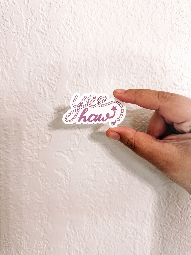 May include: A pink and white sticker with the text "yee haw" written in a rope-like font. The rope is tied into a knot at the end of the word "haw" and has a small star on it.