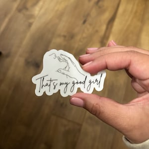 May include: A white sticker with the phrase "That's my good girl" in a cursive font. The sticker features a line drawing of a hand holding a cigarette. The sticker is held by a person.