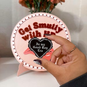 以下が含まれることがあります： 縁に有刺鉄線が巻かれたハート型の黒いエナメルピン。ピンにはゴシック体で「In my Good Boy Era」と書かれています。