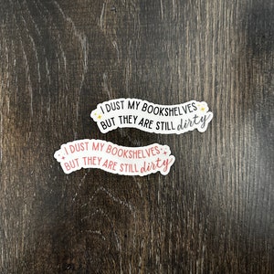 May include: Two stickers with the text "I dust my bookshelves but they are still dirty" in black and pink lettering with gold stars. The stickers are on a dark brown wooden surface.