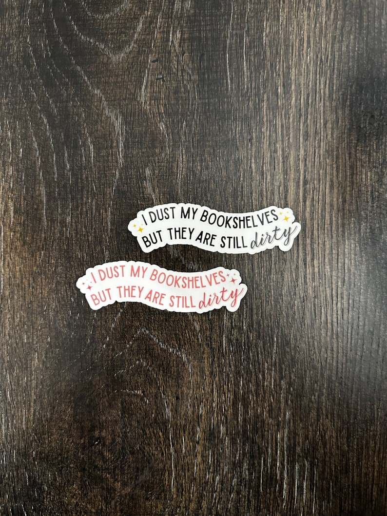 May include: Two stickers with the text "I dust my bookshelves but they are still dirty" in black and pink lettering with gold stars. The stickers are on a dark brown wooden surface.