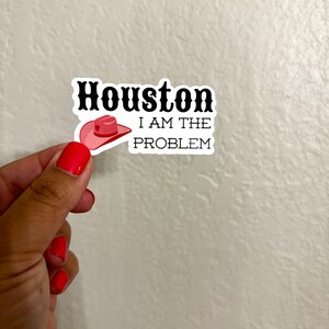 May include: A white sticker with black text that reads "Houston I am the problem". A pink cowboy hat with a red heart is above the text.