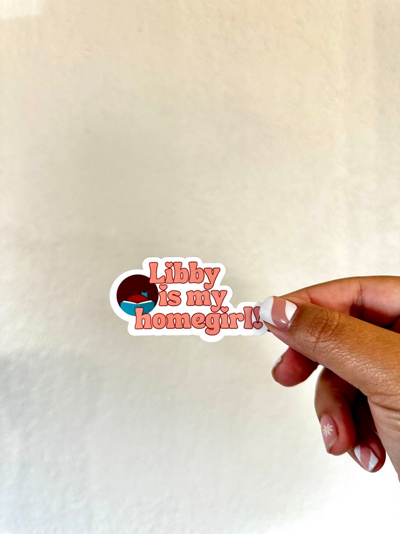 May include: A sticker with the text "Libby is my homegirl!" in pink and white, with a small graphic of a fish in a bowl. The sticker is held by a hand against a white background.