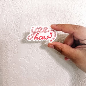 May include: A pink "yee haw" sticker with a rope design. The sticker has a small star at the end of the rope.