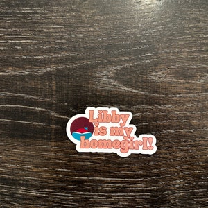 May include: A sticker with the text "Libby is my homegirl!" in a retro font. The sticker has a white border and a design of a circle with a blue and red shape inside. The background is a dark wood grain.