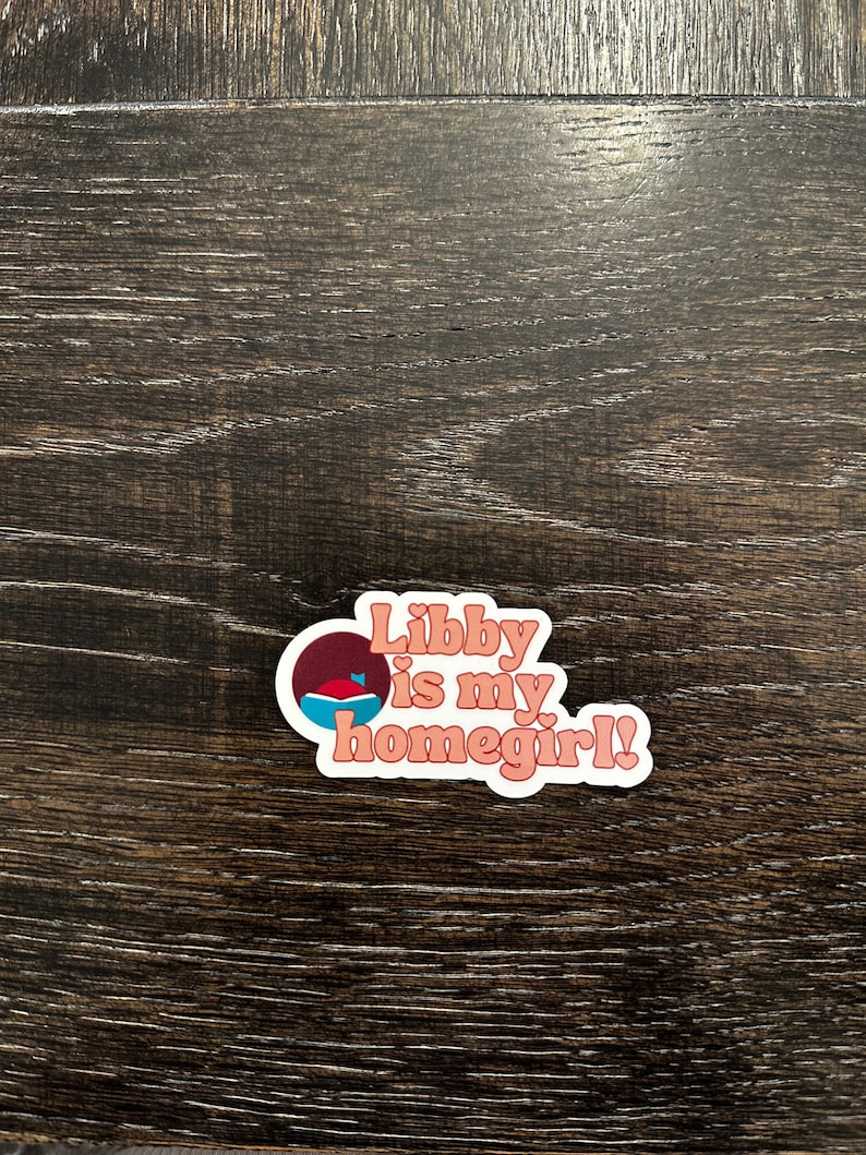 May include: A sticker with the text "Libby is my homegirl!" in a retro font. The sticker has a white border and a design of a circle with a blue and red shape inside. The background is a dark wood grain.