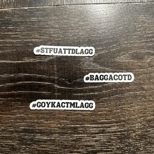 May include: Three white stickers with black text on a dark wood surface. The stickers read "#STFUATTDLAGG", "#BAGGACOTD", and "#GOYKACTMLAGG".