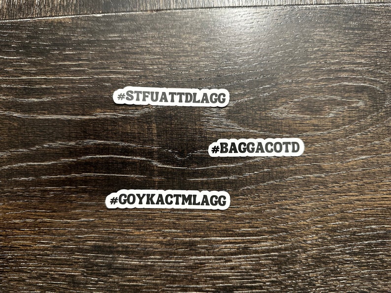 May include: Three white stickers with black text on a dark wood surface. The stickers read "#STFUATTDLAGG", "#BAGGACOTD", and "#GOYKACTMLAGG".