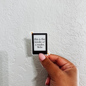 May include: A sticker shaped like a Kindle e-reader, featuring the text "this is the kindle of a reader, Bella". The sticker is held by a hand with pink nail polish against a white textured wall.