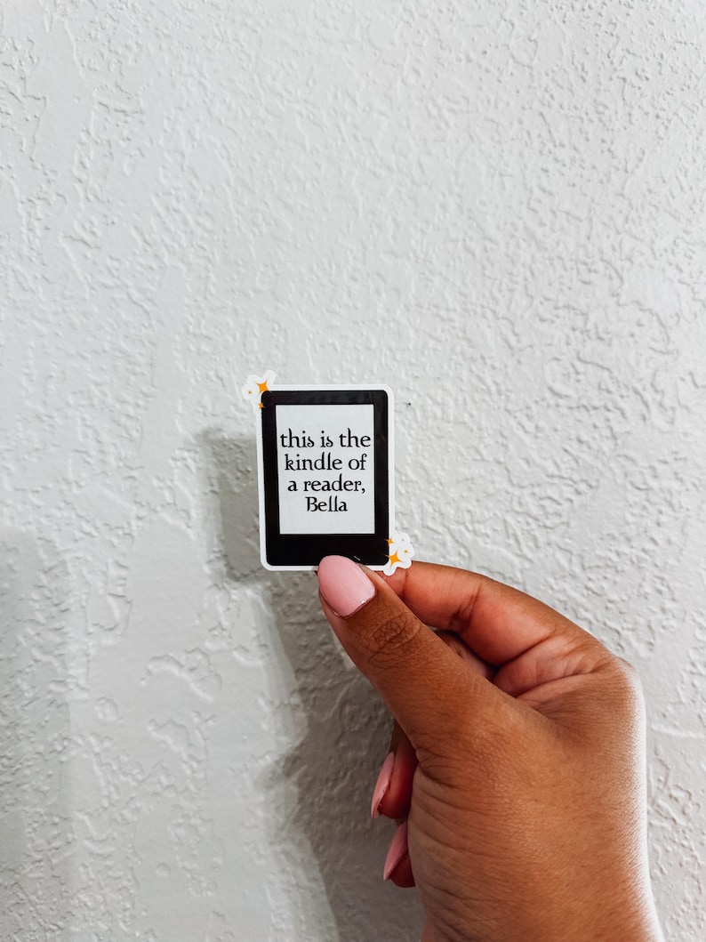 May include: A sticker shaped like a Kindle e-reader, featuring the text "this is the kindle of a reader, Bella". The sticker is held by a hand with pink nail polish against a white textured wall.