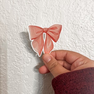 K&ouml;nnte beinhalten: Ein rosa Schleifenaufkleber mit Satin-Finish. Der Aufkleber befindet sich auf einem wei&szlig;en, strukturierten Hintergrund.