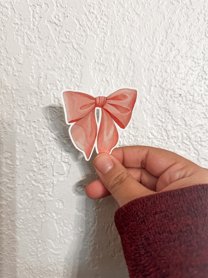 K&ouml;nnte beinhalten: Ein rosa Schleifenaufkleber mit Satin-Finish. Der Aufkleber befindet sich auf einem wei&szlig;en, strukturierten Hintergrund.