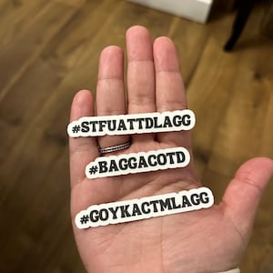 May include: Three white stickers with black text on a wooden surface. The stickers read "#STFUATTDLAGG", "#BAGGACOTD", and "#GOYKACTMLAGG".