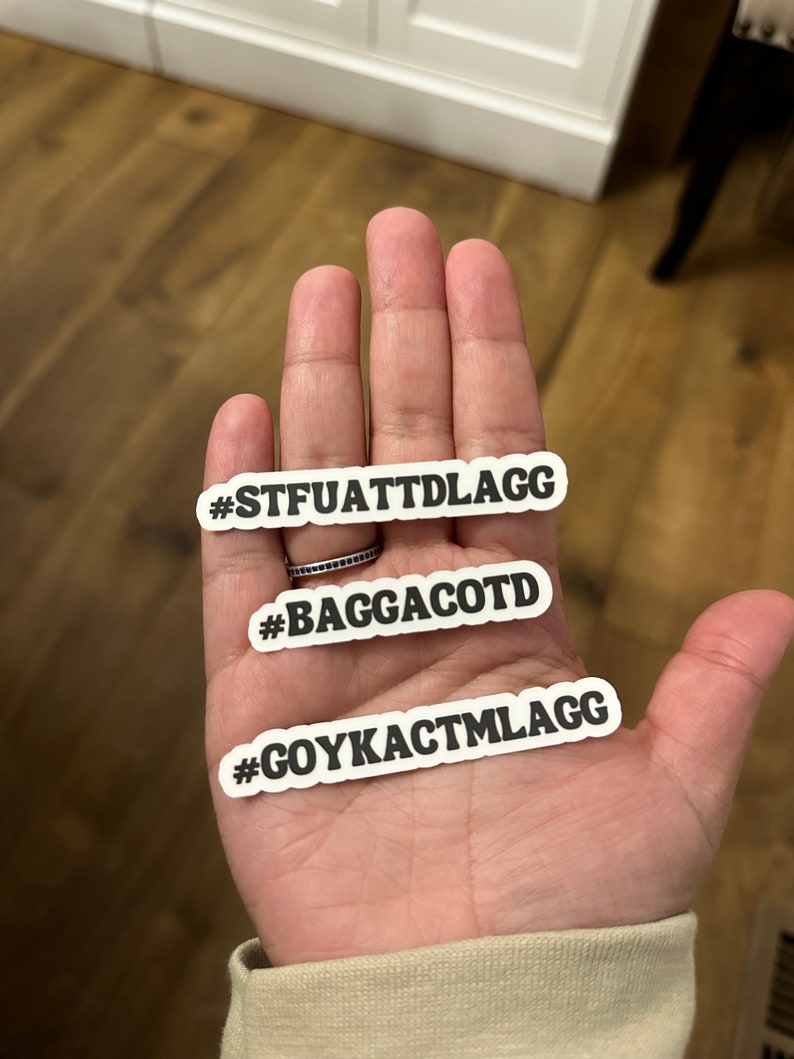 May include: Three white stickers with black text on a wooden surface. The stickers read "#STFUATTDLAGG", "#BAGGACOTD", and "#GOYKACTMLAGG".