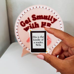 May include: A hand holding a rectangular sticker with rounded corners. The sticker has a black and white design and the text "this is the kindle of a reader, Bella." Behind it is a pink circular sign with the text "Get Smutty With HR."