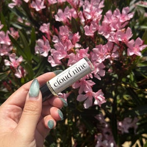 May include: A clear glass essential oil roller bottle with a black cap is held in front of a background of pink flowers. The label reads "cloud nine" with the words "evoke a floating, deeply relaxed state."
