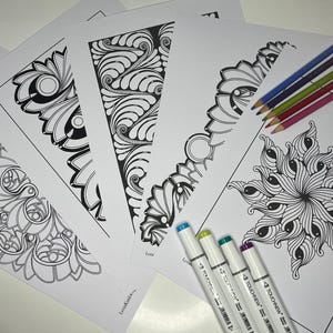 Op de afbeelding: Vier zwart-witte kleurplaten met ingewikkelde ontwerpen. De pagina's zijn in een waaier vorm gerangschikt, met de bovenste pagina gevouwen. Vijf gekleurde stiften liggen op de pagina's.