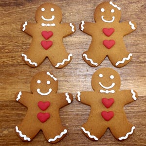 May include: Four gingerbread men biscuits with white icing details and red heart decorations. Each biscuit has a smiling face and a wavy icing border. The biscuits are arranged on a wooden surface.