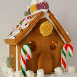 May include: A decorated gingerbread house with white icing, colourful sweets, and marshmallows. The roof is adorned with various sweets, and candy canes are placed on the sides. The house features a door and two round windows.