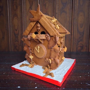 May include: A gingerbread house with a bird on top, decorated with brown icing and edible flowers. The house has a clock face and is set on a white base with a red border. A festive, edible creation.