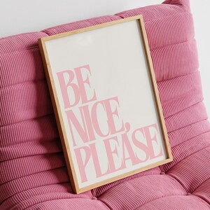 以下が含まれることがあります： ピンク色のコーデュロイラウンジチェアと額入りのプリント。プリントには、白い背景にピンク色の文字で「BE NICE, PLEASE」と書かれています。フレームは明るい木の色です。