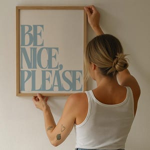 以下が含まれることがあります： 「BE NICE, PLEASE」の文字が水色のフレーム入りプリント。プリントは薄茶色の木製フレームに入っています。白い壁にフレーム入りプリントを持っている人。