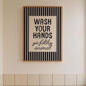 May include: A bathroom interior with a framed print saying "WASH YOUR HANDS ya filthy animal." The print has a black and white striped border. A wooden stool holds a bottle and brush. A white bathtub and a window are also visible.