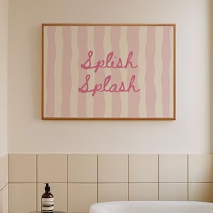 May include: A bathroom interior with a framed print reading "Splish Splash" in pink script on a pink and beige striped background. A wooden stool holds a bottle and brush, and a white towel hangs on the edge of a white bathtub.