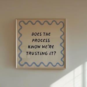May include: A square wall art print in a light-colored frame featuring a blue wavy border and the black text "DOES THE PROCESS KNOW WE'RE TRUSTING IT?" centered on a cream background, displayed above a wooden slatted media console cabinet.