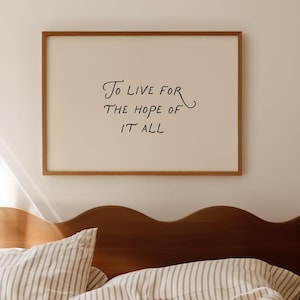 以下が含まれることがあります： 「To Live For The Hope Of It All」というテキストが黒い筆記体で書かれた額入りのプリント。フレームは温かみのある茶色で、背景は明るいクリーム色です。プリントはベッドの上に掛けられています。
