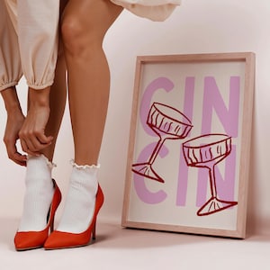 Puede incluir: Un cuadro enmarcado con la palabra "GIN" en rosa y dos copas de c&oacute;ctel rojas. La imagen est&aacute; apoyada contra una pared. La persona lleva tacones altos naranjas y calcetines blancos.