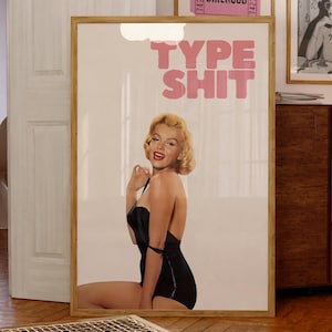 May include: A framed print featuring Marilyn Monroe in a black swimsuit with the text "TYPE SHIT" in pink. Two framed pink "GIRLHOOD" tickets and a framed newspaper print are also visible. The room has wooden floors and a wooden cabinet.