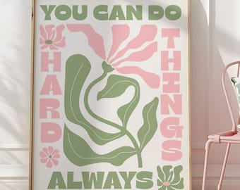 May include: A framed art print with a botanical design and the words "YOU CAN DO HARD THINGS ALWAYS" in green and pink. The print is displayed on a wooden floor next to a pink chair with a pillow.