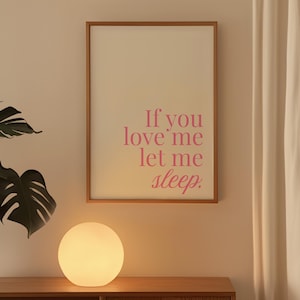 以下が含まれることがあります： ピンク色の文字で「If you love me let me sleep」と書かれた額入りのプリント。ベージュの壁に掛けられており、木製のキャビネットの上にあります。キャビネットには丸く光るランプが置かれています。