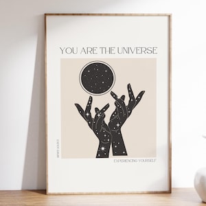 以下が含まれることがあります： 「You are the universe」と「Experiencing yourself」というテキストが書かれた、ベージュとブラックのミニマルなポスター。ポスターには、星が描かれた2つの手が、星が描かれた黒い円に向かって伸びている様子が描かれている。