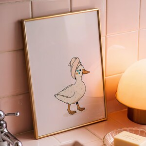 May include: A framed print featuring a cartoon duck wearing a towel on its head. The duck is white with brown feet and a yellow beak. The print is in a gold frame, and the background is a pink tiled wall. A pink sink, soap dish, and lamp are also visible.