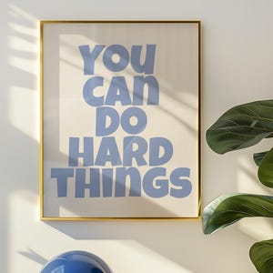 Peut inclure: Une affiche encadrée avec le texte "YOU CAN DO HARD THINGS" en bleu clair. Une lampe de table bleue avec une base dorée est posée sur une surface blanche. Une plante en pot avec des feuilles vertes est à droite.