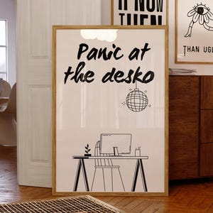 May include: A framed art print with the text "Panic at the desko" in a handwritten style, with a disco ball illustration. The print also features a desk with a computer and chair. Two other framed prints are visible in the background.