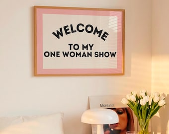 Könnte beinhalten: Ein gerahmter Druck mit dem Text "WELCOME TO MY ONE WOMAN SHOW" in Schwarz auf weißem Hintergrund, umrahmt in Rosa und Holz. Das Bild befindet sich über einem Bett mit weißer Bettwäsche und einem Nachttisch aus Holz mit einer Lampe und Blumen.