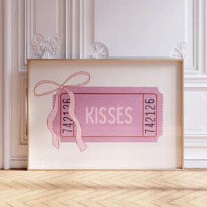 May include: A pink ticket-style art print with the word 'Kisses' in the center. The ticket has a pink bow and ribbon at the top left. The numbers '742126' are printed on both sides of the ticket. The print is framed in a light brown frame and displayed against a white wall with decorative molding.