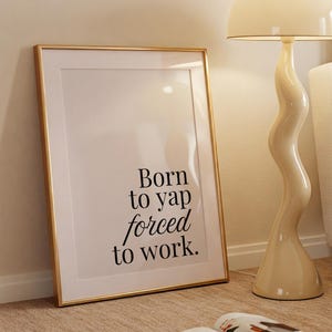 May include: A cream-colored floor lamp with a wavy stem and a dome-shaped shade. A framed print with the text "Born to yap forced to work" leans against the wall. A magazine is open on the floor.