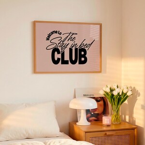 Op de afbeelding: Een slaapkamer met een bed met witte lakens en kussens. Een houten nachtkastje toont een lamp, een vaas met witte tulpen en een ingelijste print met de tekst "Welcome to The Stay in bed CLUB".