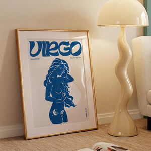 K&ouml;nnte beinhalten: Ein gerahmtes Poster mit einer blauen Illustration einer Frau, die das Sternzeichen Jungfrau darstellt. Das Poster hat einen goldenen Rahmen und liegt auf einem beigefarbenen Teppich. Der Text auf dem Poster lautet "Virgo" und "23. August - 22. September".