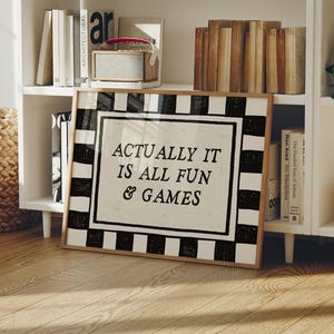 May include: A framed sign with the text "ACTUALLY IT IS ALL FUN & GAMES" in a black and white checkered border. The sign is displayed on a wooden floor in front of a white bookshelf with books and decorative items.