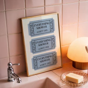 May include: A framed print with the text "EVERYTHING SHOWER ADMIT ONE" on three blue tickets. The bathroom scene includes a pink sink, a gold-rimmed lamp, a toothbrush, and a soap dish.