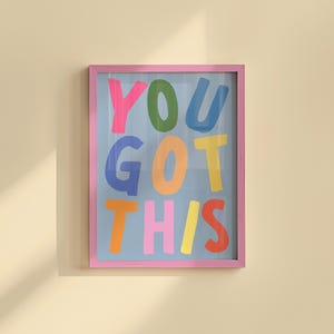 May include: A framed art print with the words "YOU GOT THIS" in colorful, playful lettering. The letters are in pink, green, blue, orange, yellow, and light pink against a light blue background, framed in a pink border.