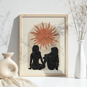 以下が含まれることがあります： 2 人のシルエットが背中合わせに座り、大きなオレンジ色の太陽と光線を仰ぎ見ている、ミニマルなデザインの額装されたプリント。背景は薄いベージュ色です。