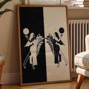 以下が含まれることがあります： 背景に葉っぱのデザインが施された、2 人の人物が球体を持っている白黒の額装されたプリント。プリントは、ソファと植物のある部屋の壁に飾られています。