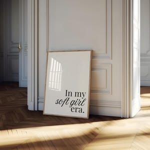 Puede incluir: Un estampado enmarcado con el texto "In my soft girl era". El estampado est&aacute; en un marco de madera clara y se apoya contra una pared blanca. La habitaci&oacute;n tiene un suelo de madera y una puerta al fondo.