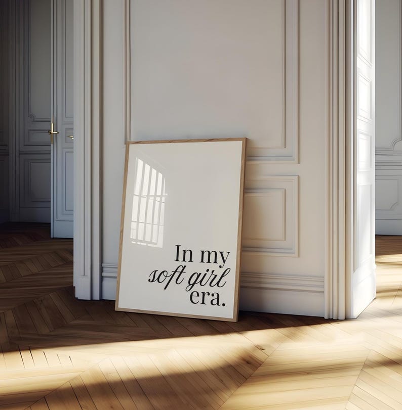 Puede incluir: Un estampado enmarcado con el texto "In my soft girl era". El estampado est&aacute; en un marco de madera clara y se apoya contra una pared blanca. La habitaci&oacute;n tiene un suelo de madera y una puerta al fondo.
